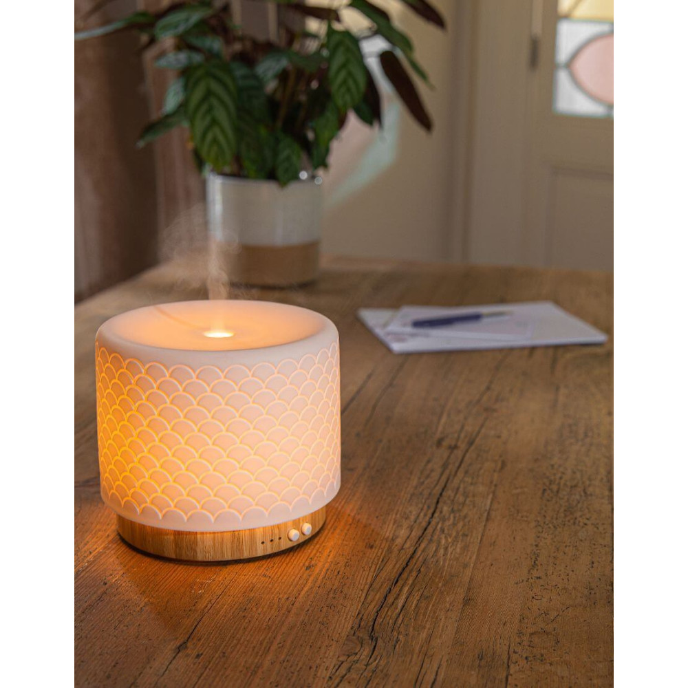 Petit diffuseur/lampe d’ambiance en bois avec abat-jour blanc à motif écailles, diffusant une lumière chaude sur une table en bois. Plant et documents flous en arrière-plan.